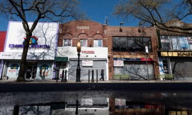 A woman wearing a mask walks past closed storefronts in the Astoria neighborhood of Queens