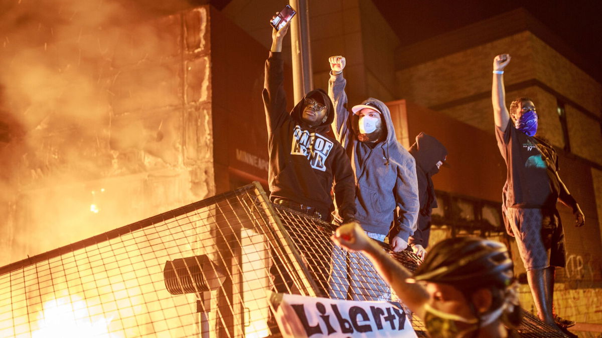 <i>Kerem Yucel/AFP via Getty Images</i><br/>Protesters hold up their fists as flames rise behind them in front of the Third Police Precinct on May 28