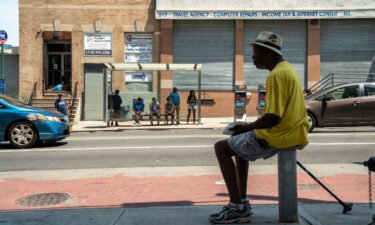 A man sits in the shade a people wait at a bus stop in the Bronx borough of New York