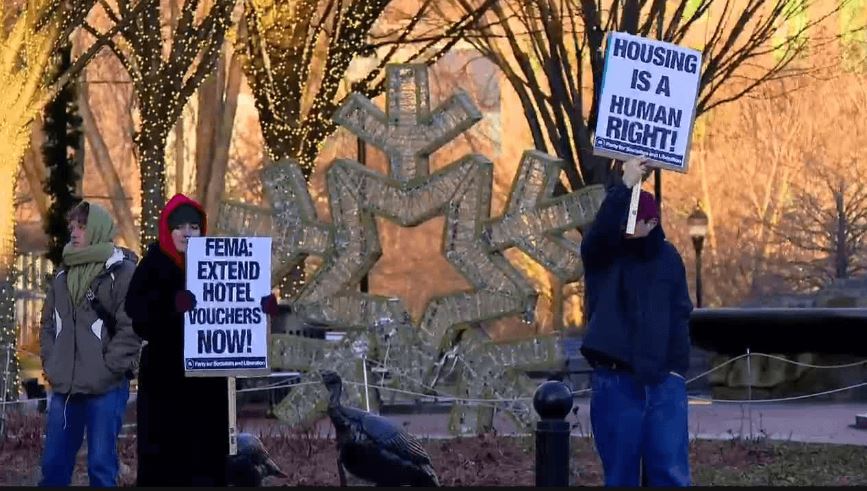 <i>WLOS via CNN Newsource</i><br/>Demonstrator holds a sign in downtown Asheville calling on FEMA to extend temporary housing assistance to victims of Helene currently staying in hotels after the storm.